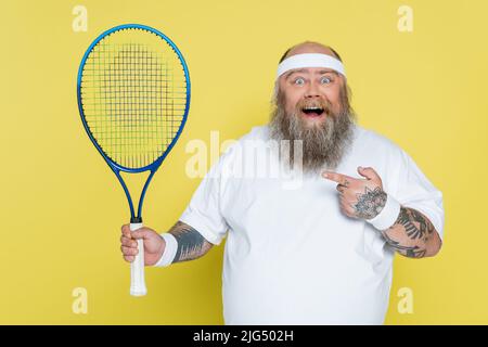 homme de grande taille stupéfié pointant vers une raquette de tennis et regardant un appareil photo isolé sur le jaune Banque D'Images