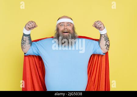 sportif bald et barbu en surpoids dans un manteau de super-héros montrant le pouvoir isolé sur le jaune Banque D'Images