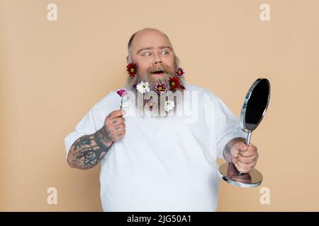 homme de taille plus stupéfait avec des fleurs dans la barbe regardant dans le miroir isolé sur beige Banque D'Images