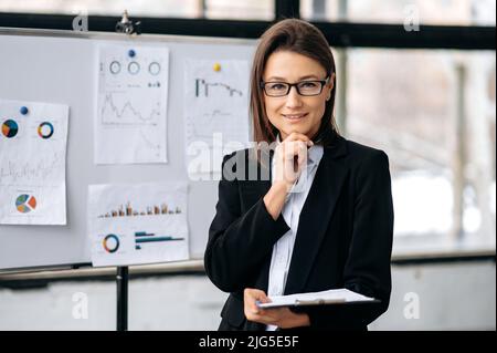 Portrait d'une femme d'affaires caucasienne intelligente et confiante avec des lunettes, dans des vêtements d'affaires, entrepreneur, pdg de la société, se tient dans le bureau près du tableau de marqueur, regarde la caméra, sourit Banque D'Images