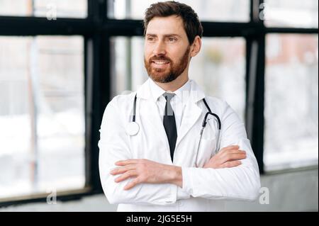 Professionnel réussi fier homme caucasien médecin, médecine générale, en uniforme médical, avec un stéthoscope sur ses épaules, se tient avec ses bras croisés à l'hôpital, regarde loin, sourit Banque D'Images