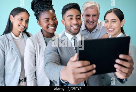 En prenons quelques autres. Prise de vue en studio d'un groupe diversifié de professionnels utilisant une tablette pour prendre des selfies sur fond bleu. Banque D'Images