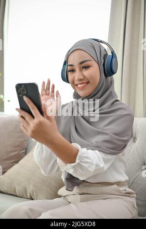 Belle femme musulmane asiatique du millénaire ayant un appel vidéo de ses amis dans le salon. Banque D'Images