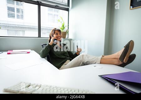 Jeune femme d'affaires asiatique souriante parlant sur un téléphone portable tout en se relaxant au bureau Banque D'Images