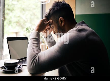 .. Un jeune homme de course mixte stressé assis à une table dans un café, buvant un café et utilisant un ordinateur portable seul. Malheureux homme d'affaires hispanique assis dans un Banque D'Images