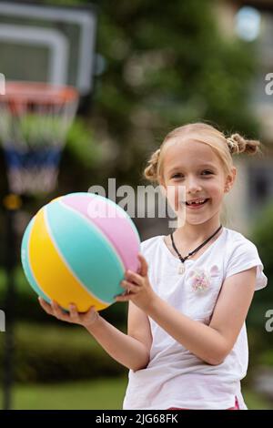 Portrait de style de vie candid de bonne joyeuse caucasienne petite fille avec des cheveux blonds sept ans tenant le ballon de jeu pour le basket-ball extérieur à chaud Banque D'Images