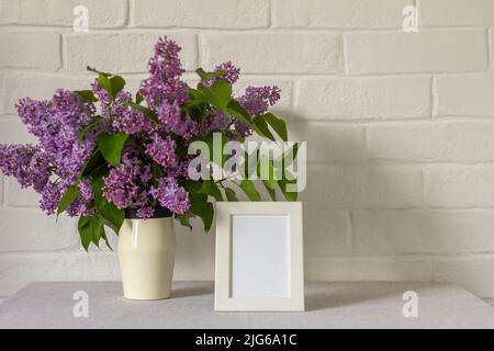 Maquette d'un cadre photo blanc sur la table et d'un bouquet de lilas dans un vase.fond de mur blanc. Intérieur scandinave Banque D'Images