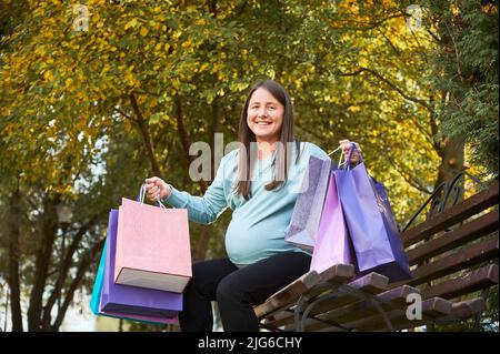 Une femme enceinte gaie souriant à l'appareil photo après avoir fait du shopping pendant la journée pendant l'attente DE BÉBÉ. Bonne femme avec des sacs en papier appréciant les loisirs pour se préparer à l'apparence de tout-petit. Banque D'Images
