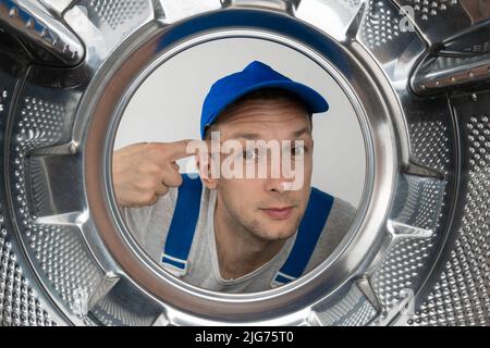 Le réparateur mâle en uniforme regarde dans le tambour de la machine à laver et tourne son doigt sur le temple, photo de l'intérieur. Banque D'Images