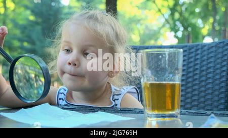 La petite fille regarde dans l'objectif autour. Gros plan d'une fille blonde étudiant le monde autour d'elle la regardant à travers la loupe tout en étant assise Banque D'Images