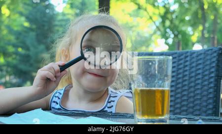 La petite fille regarde dans l'objectif autour. Gros plan d'une fille blonde étudiant le monde autour d'elle la regardant à travers la loupe tout en étant assise Banque D'Images