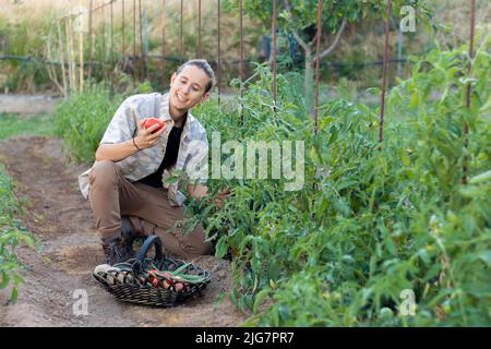 Jeune homme heureux et souriant d'avoir recueilli sa première tomate rouge et fraîche de son verger de jardin naturel Banque D'Images