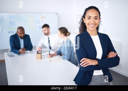 Portrait d'une femme d'affaires souriante et confiante de la course mixte, penchée sur un bureau dans une salle de réunion avec les bras repliés. Divers groupes d'hommes d'affaires en réunion et Banque D'Images