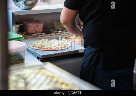 Un homme méconnu qui met du fromage sur des pizzas dans une cuisine de restaurant. Banque D'Images