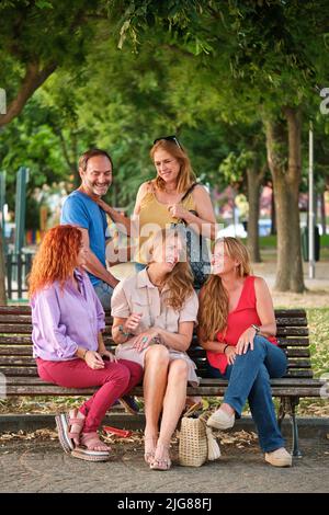 Cinq adultes matures parlant et riant assis sur un banc dans un parc. Banque D'Images