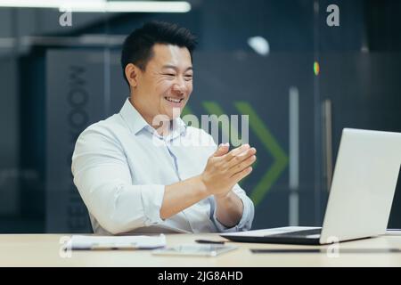 Un homme d'affaires asiatique satisfait regardant l'écran d'ordinateur portable, célébrant la victoire et le triomphe, se frottant les mains ensemble, geste de succès attendu. Banque D'Images