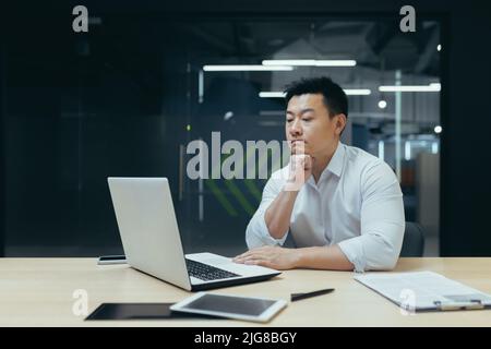Homme d'affaires asiatique pensif travaillant dans un bureau moderne, homme pensant à la décision, directeur expérimenté avec ordinateur portable. Banque D'Images