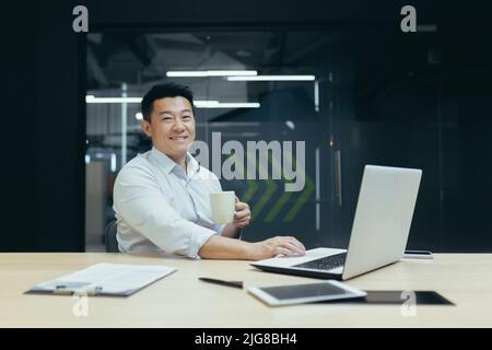 Portrait d'un patron asiatique réussi, homme avec une tasse de boisson chaude travaillant dans un bureau moderne, souriant et regardant l'appareil photo. Banque D'Images