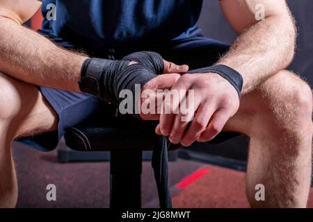 Corps court de jeune artiste martial mixte fort qui s'assoit sur le canapé et enveloppe le bandage élastique pour fixer son poignet pour le combat ouvert de poing. Banque D'Images