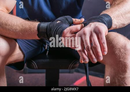 Corps court de jeune artiste martial mixte fort qui s'assoit sur le canapé et enveloppe le bandage élastique pour fixer son poignet pour le combat ouvert de poing. Banque D'Images