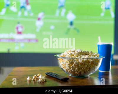 L'ensemble traditionnel d'un fan de sport - pop-corn dans un bol en verre, une boisson gazeuse dans un verre et une télécommande sur la table avec la toile de fond Banque D'Images