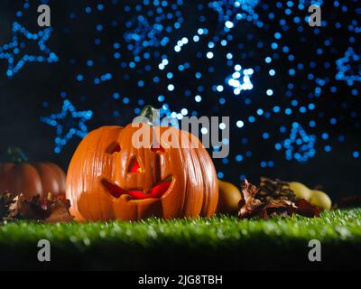 Composition mystique d'Halloween. Citrouille d'halloween souriante avec visage sculpté illuminé, fruits d'automne, cônes sur une pelouse verte contre un bleu chatoyant Banque D'Images