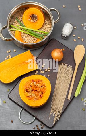 Potiron et nouilles, cuillère en bois et oignon sur planche à découper. Pois chiches, citrouille et gousses vertes dans une casserole. Pose à plat. Arrière-plan gris. Banque D'Images