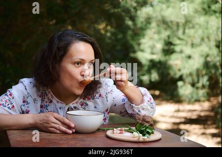 Agréable femme multiethnique dans la campagne de l'Ukraine tente le plat national ukrainien. Soupe traditionnelle de betteraves au borsch servie avec des oignons verts, ga Banque D'Images