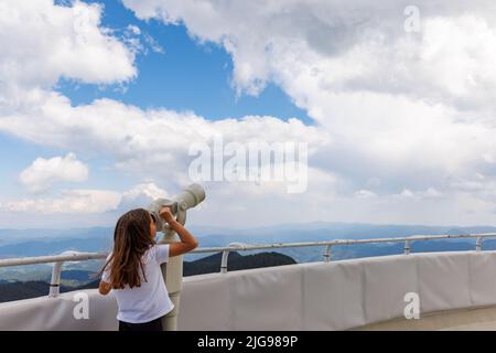 Petite fille curieuse regardant les paysages de la vallée de la forêt de montagnes de Rhodope et bleu clair ciel nuageux à travers le télescope métallique moderne, on Banque D'Images