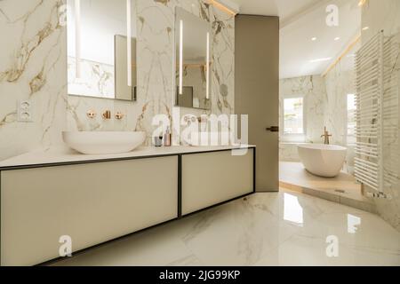 Salle de bains avec grand meuble suspendu avec évier en porcelaine séparé, deux miroirs assortis, murs en marbre, baignoire blanche et radiateur blanc Banque D'Images