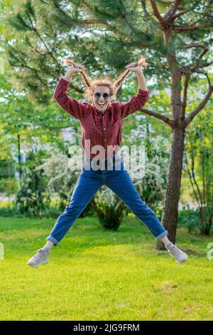 Une femme en riant sauta dans l'air en tenant ses deux queues de porc dans le parc vert de printemps. Cheveux forts. Banque D'Images