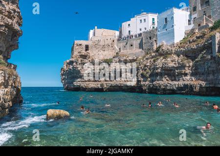 Belle vue de Polignano a Mare avec maisons en pierre blanche, falaise, plage, mer bleue et touristes, baigneurs, entouré par la nature méditerranéenne Banque D'Images