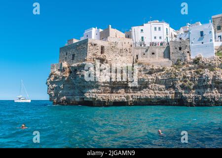 Belle vue de Polignano a Mare avec maisons en pierre blanche, falaise, plage, mer bleue et touristes, baigneurs, entouré par la nature méditerranéenne Banque D'Images