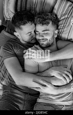 Un cliché vertical d'un couple homosexuel hispanique et caucasien masculin qui se couche dans un lit en cuddling heureux en niveaux de gris Banque D'Images