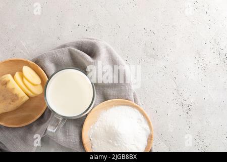 Tasse en verre avec lait de pomme de terre, poudre de pomme de terre et tubercule coupé en plaques de bois sur serviette grise sur béton. Lait de légumes en poudre. Remplacement du lait sans lactose Banque D'Images