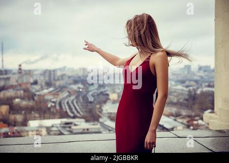 Fille mince en robe rouge est debout au-dessus de gratte-ciel, haut au-dessus de la mégacité s'étendant derrière elle jusqu'à l'horizon. Elle pointe avec 1 mains Banque D'Images