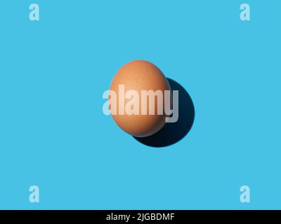 Un œuf de poulet brun se trouve au centre sur un fond bleu vif. Lumière vive, ombre profonde. Gros plan, espace de copie, vue de dessus. Banque D'Images