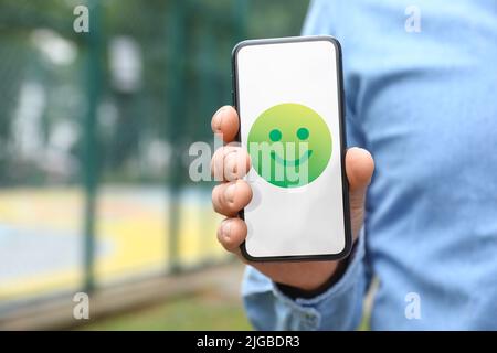 Homme tenant un téléphone avec une émoticône heureuse à l'écran en plein air, en gros plan Banque D'Images