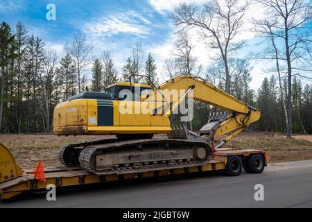 Chargeuse-pelleteuse jaune d'occasion sur une remorque à plateau, sur un chantier de construction, dans la lumière du soir avec des arbres en arrière-plan. Banque D'Images