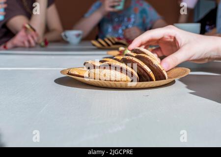 Une assiette en papier marron avec des biscuits enrobés de chocolat est posée sur un comptoir de cuisine beige clair. Les gens boivent du thé. Une main prend le cookie de l'assiette. Banque D'Images