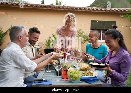 Une femme de race blanche assez souriante d'âge moyen qui met une assiette avec de la nourriture sur la table à manger lors d'une fête avec des amis heureux dans l'arrière-cour. Banque D'Images