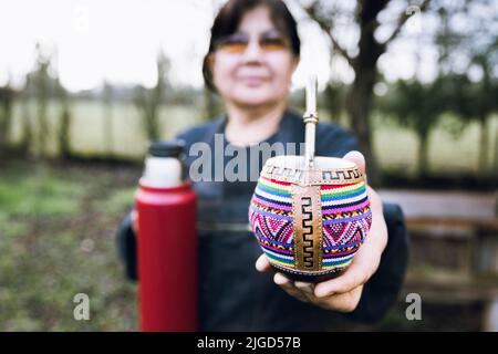 Femme latine au service de son compagnon avec un thermo rouge, dans un compagnon ethnique coloré. Banque D'Images