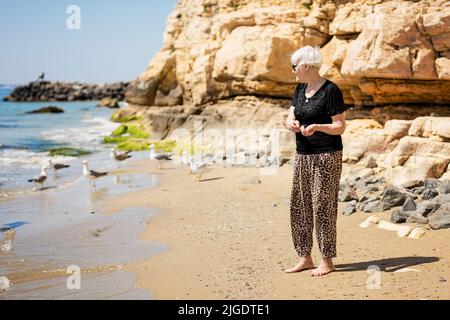 Une femme âgée en lunettes de soleil foncées nourrit des mouettes sur la plage Banque D'Images