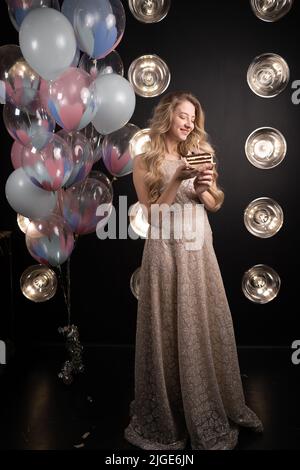 Femme heureuse avec ballons. Jeune fille debout dans une robe avec un morceau de gâteau d'anniversaire sur un fond noir. Rires merrly, émotions de joie et Banque D'Images