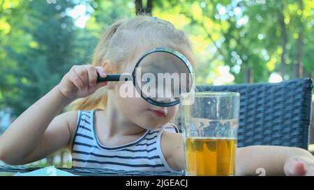 La petite fille regarde dans l'objectif autour. Gros plan d'une fille blonde étudiant le monde autour d'elle la regardant à travers la loupe tout en étant assise Banque D'Images