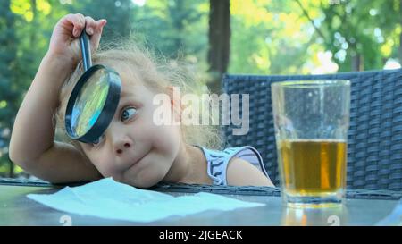 La petite fille regarde dans l'objectif autour. Gros plan d'une fille blonde étudiant le monde autour d'elle la regardant à travers la loupe tout en étant assise Banque D'Images