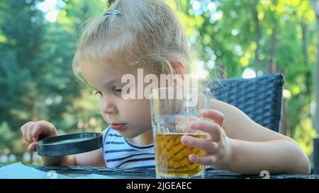 La petite fille regarde dans l'objectif autour. Gros plan d'une fille blonde étudiant le monde autour d'elle la regardant à travers la loupe tout en étant assise Banque D'Images