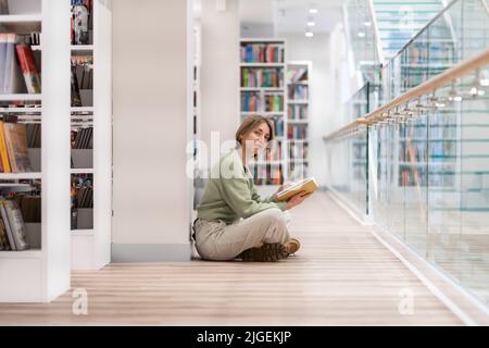 Une femme d'âge moyen liseuse assise sur le sol avec un livre entre les mains dans une bibliothèque publique moderne Banque D'Images