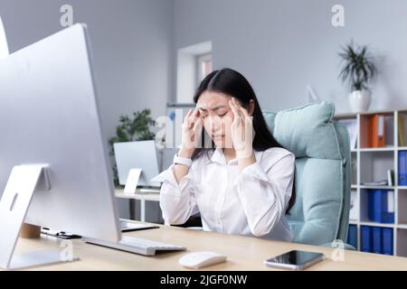 Épuisement au travail. Une jeune femme asiatique tient la tête, ressent de la douleur, grimaces. Assis dans le bureau au bureau, travaillant à l'ordinateur, épuisé. B Banque D'Images