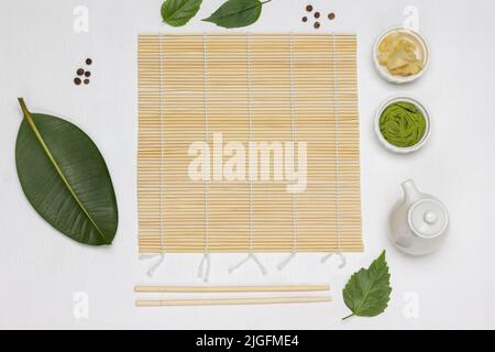 Tapis de bambou, baguettes, épices à sushis et feuilles vertes sur la table. Pose à plat. Arrière-plan blanc Banque D'Images